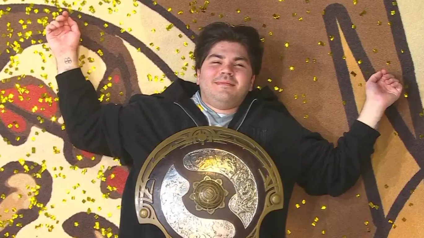 Team Spirit player lying on the floor with gold confetti, holding the Dota 2 Aegis of Champions trophy.