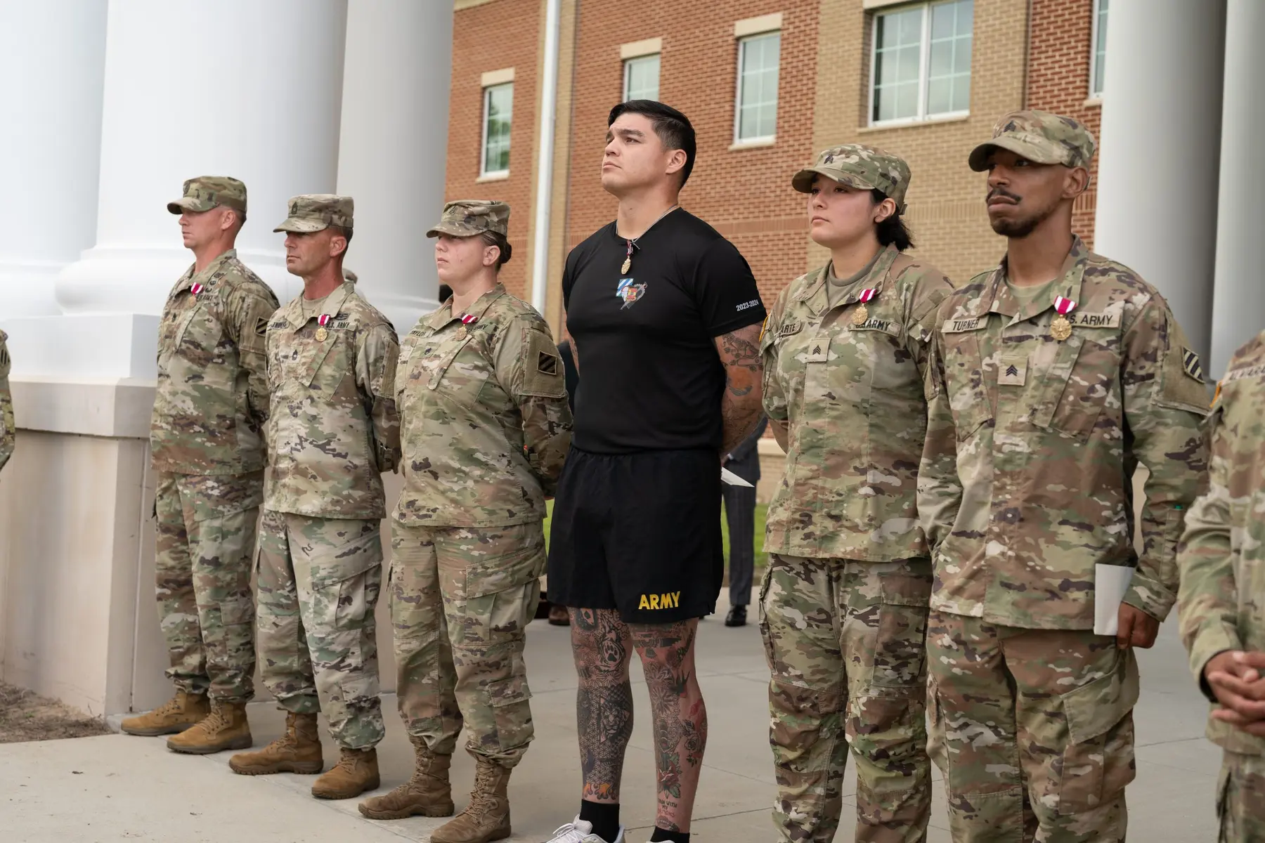 A row of US Army soldiers in OCP uniforms and one soldier in PT gear standing at attention outdoors.