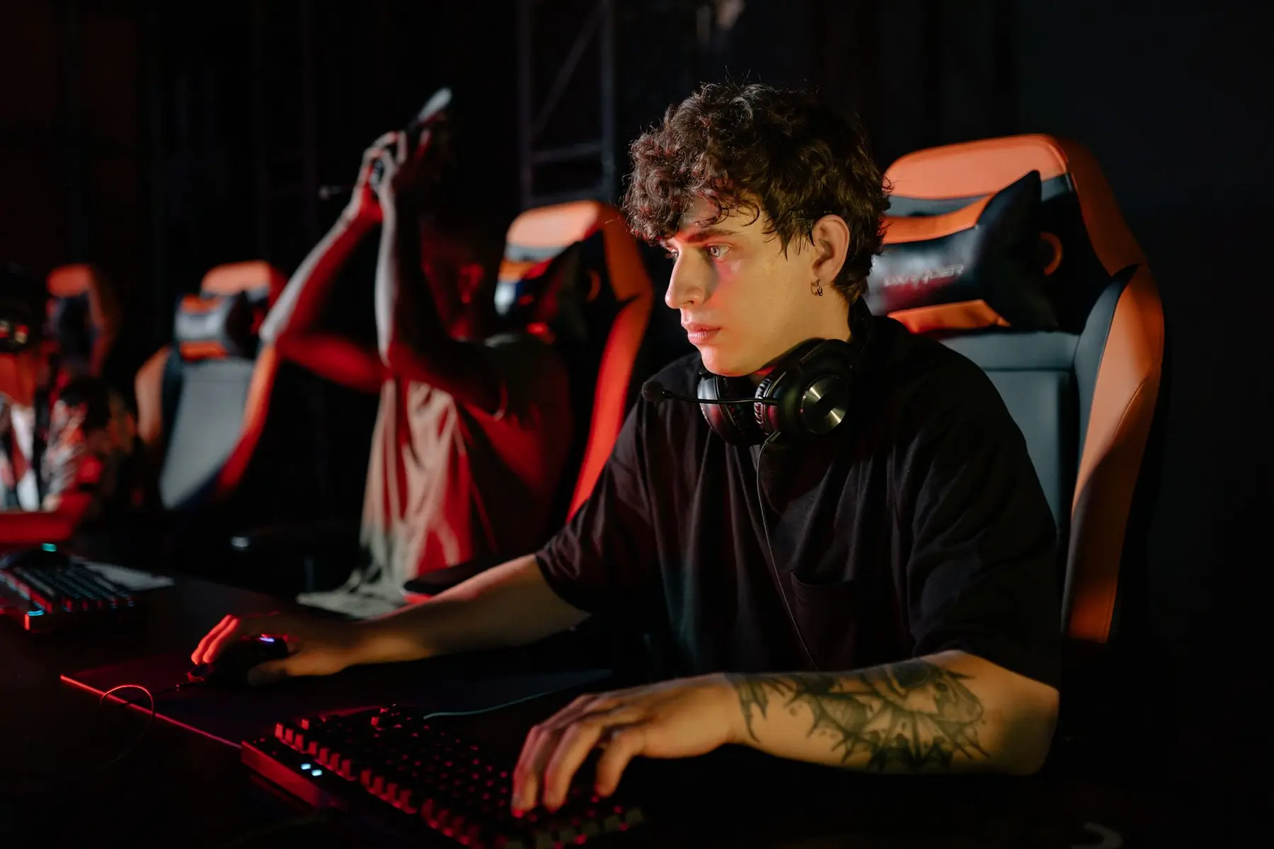 A professional gamer with curly hair and arm tattoos wearing a headset at a gaming PC setup